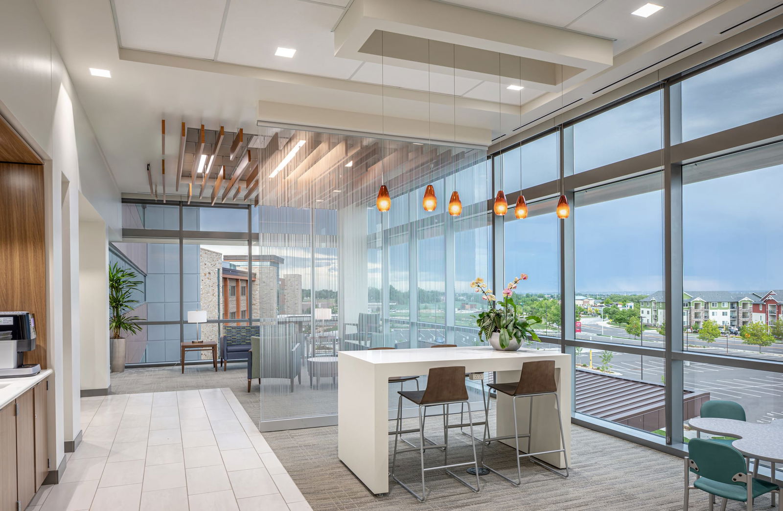 A modern healthcare waiting area with a contemporary design. The space features a mix of seating options, including blue armchairs, wooden-framed lounge chairs, and tall wooden barstools paired with a modern white freestanding table. A large window lets in natural light, showing the outside parking lot and distant tall apartments. Pendant lights with amber glass shades hang from the ceiling.