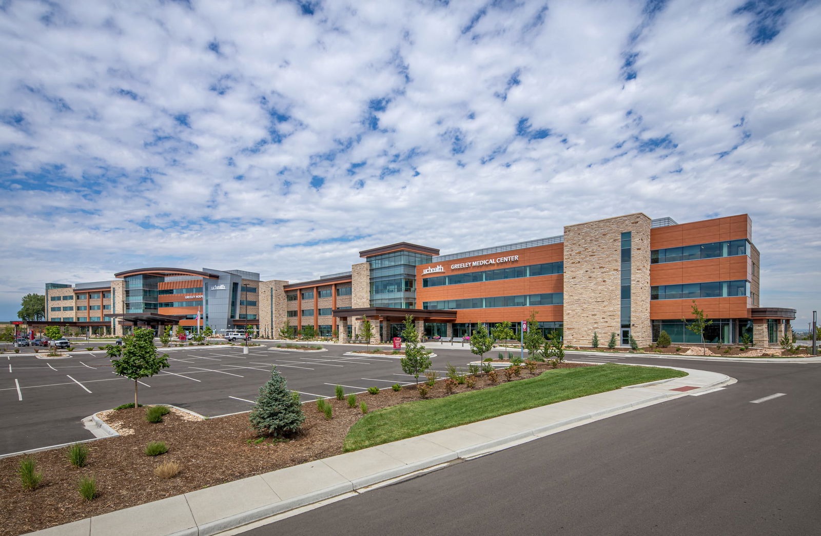 A modern hospital building labeled 'Greeley Medical Center' with the 'UCHealth' logo. The architecture features a combination of glass, metal, and stone materials, with a curved roof and large windows reflecting the sky. There are two covered drop-off areas supported by stone pillars. The surrounding landscape includes neatly trimmed grass, small trees, and decorative lighting. This image shows BSA's expertise in healthcare architecture.