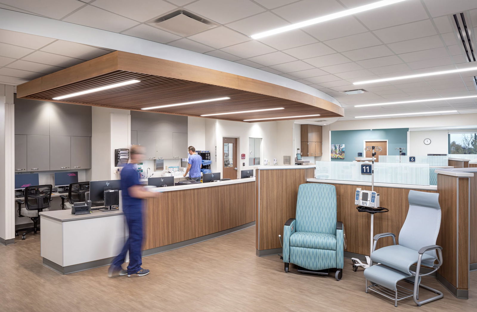 Infusion center or treatment room in the UCHealth Greeley Hospital with multiple semi-private stations. Each station has aqua blue reclining chairs with a wave pattern and is separated by wooden and glass partitions. Medical monitoring equipment is visible on small movable tables. Floor-to-ceiling windows provide natural light and views to the outdoors. The space has a curved layout with numbered stations, linear ceiling lights, and a clean, modern design with wood-tone and neutral colors.